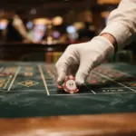 A gloved hand places a five-dollar casino chip onto a green felt roulette betting table
